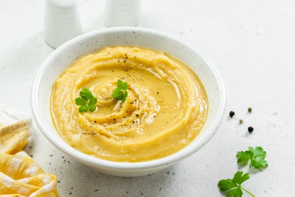 Visão superior de uma tigela branca com creme de ervilha. O creme tem uma textura aveludada, finalizado com um fio de azeite de oliva brilhante, pimenta-do-reino moída e duas pequenas folhas de salsa fresca no centro. A tigela está sobre uma superfície clara e levemente texturizada. Ao lado, aparecem grãos de pimenta inteiros espalhados e o canto de um pano de prato xadrez amarelo e branco, criando uma composição gastronômica clean e iluminada.