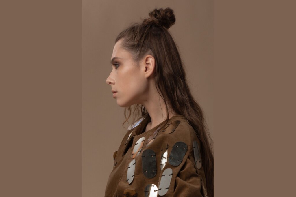 Fotografia de perfil de uma mulher com cabelos castanhos longos e ondulados, usando um penteado "half bun" (meio coque) alto e texturizado no topo da cabeça, com a metade inferior dos fios solta sobre as costas. Ela veste uma jaqueta marrom de camurça decorada com grandes aplicações ovais metálicas e reflexivas. A composição possui tons terrosos, com um fundo liso em bege escuro e iluminação suave que destaca o contorno do seu rosto e a textura do cabelo.
