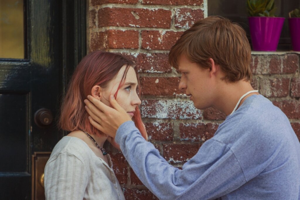 Uma cena do filme Lady Bird com dois jovens de pele clara em um momento íntimo. À esquerda, a personagem Lady Bird (Saoirse Ronan), com cabelos curtos de tom rosa avermelhado, olha fixamente para o jovem à sua frente. Ele está à direita, veste uma camiseta cinza de manga longa e segura delicadamente o rosto dela com a mão esquerda. Eles estão em frente a uma parede de tijolos vermelhos rústicos, ao lado de uma porta de madeira escura.
