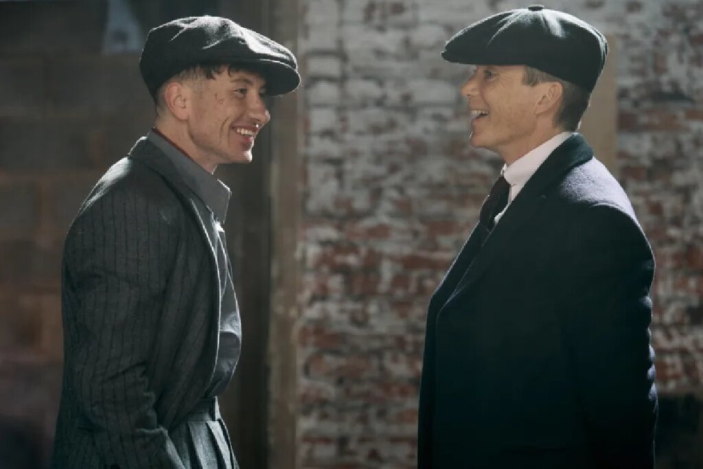 Dois homens de pele clara, interpretados pelos atores Barry Keoghan e Cillian Murphy, estão de perfil, um de frente para o outro, sorrindo e conversando. Ambos vestem figurinos de época da série Peaky Blinders, incluindo sobretudos escuros e as características boinas de lã (newsboy caps). O fundo é uma parede de tijolos rústica e desfocada.