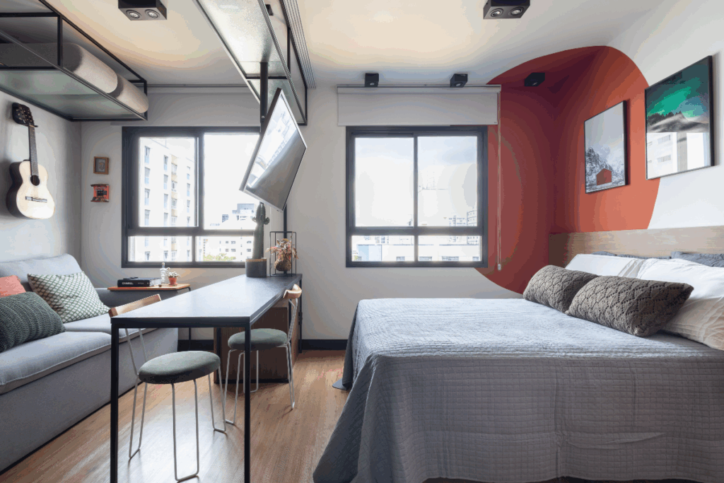 Quarto com sala integrada, móveis em tons de cinza, com TV presa em prateleira e dividindo os ambientes, a parede é branca com um círculo laranja em destaque com quadros na parede e duas janelas ao fundo