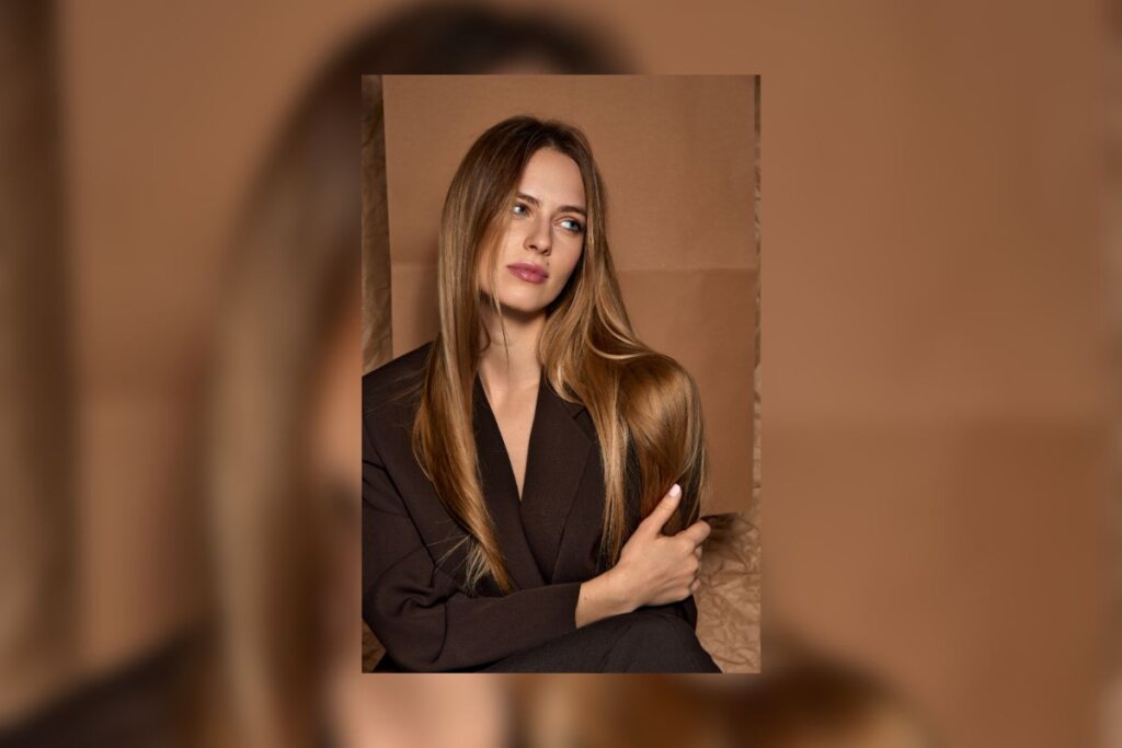 Mulher, com cabelo loiro e longo, usando blazer preto em frente a um fundo bege