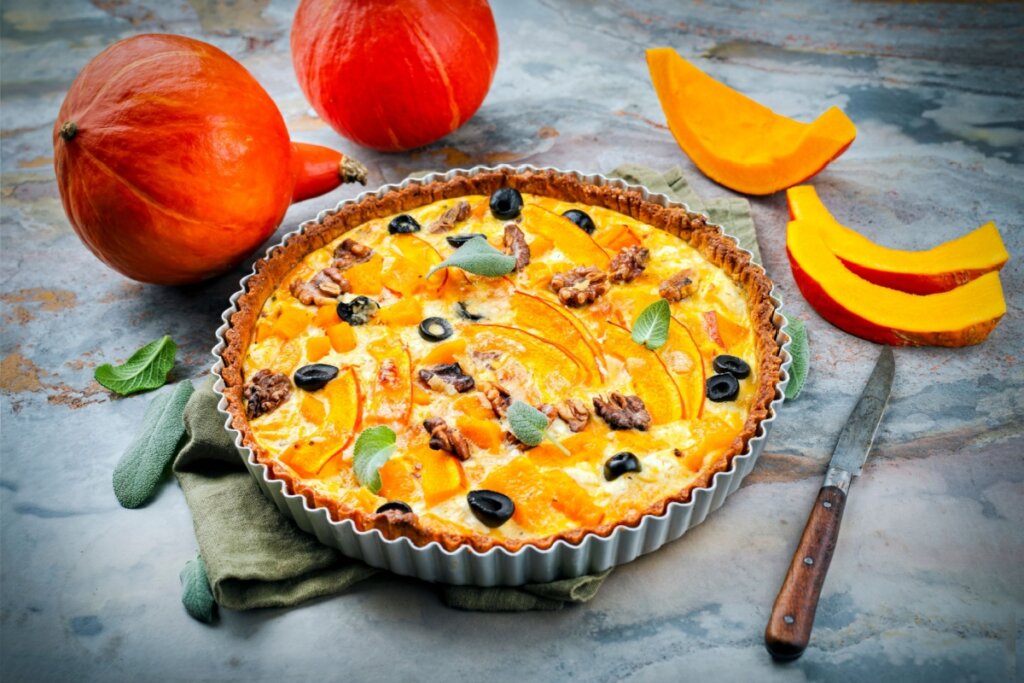 Quiche de abóbora com nozes e azeitonas em cima de uma mesa cinza com pedaços de abóbora