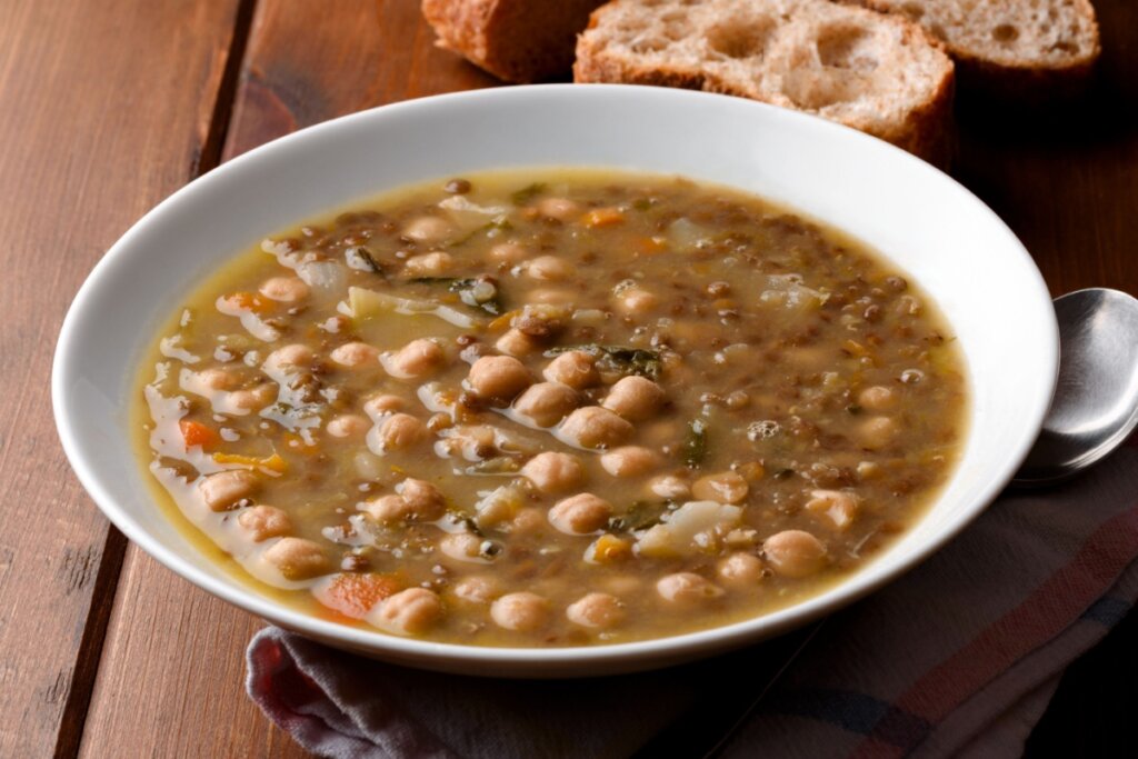 Fotografia em plano superior de uma tigela de cerâmica rústica contendo uma sopa de grãos (lentilhas e grão-de-bico) em um caldo espesso e dourado, decorada com ervas frescas e um fio de azeite. A tigela está sobre um pano de prato listrado em uma superfície de madeira. Ao lado, encontram-se fatias de pão integral, uma colher de metal e pequenos recipientes com temperos e grãos crus, criando uma composição culinária acolhedora e nutritiva.