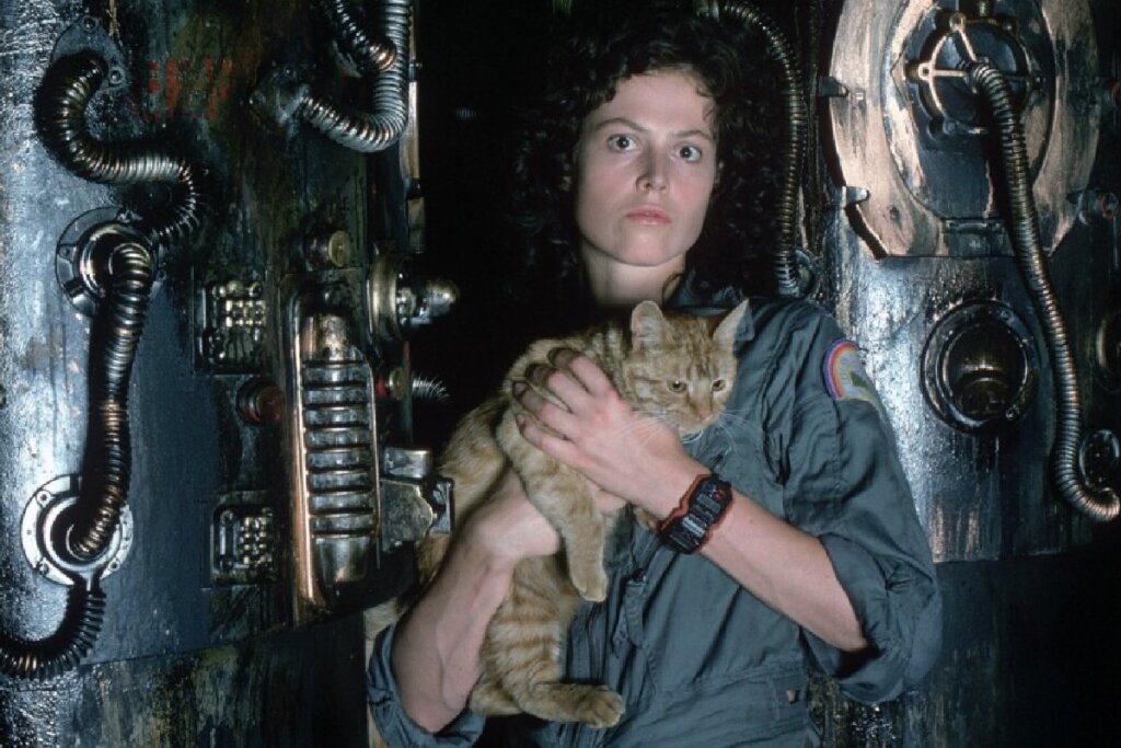 A personagem Ellen Ripley é mostrada do peito para cima, segurando um gato alaranjado contra o corpo. Ela veste um macacão de voo cinza escuro com um emblema colorido no ombro e um relógio digital preto robusto no pulso. Ripley tem cabelos escuros e cacheados e uma expressão de alerta ou medo nos olhos. O fundo é composto pelas paredes metálicas, escuras e cheias de tubulações e fiações industriais da nave Nostromo.