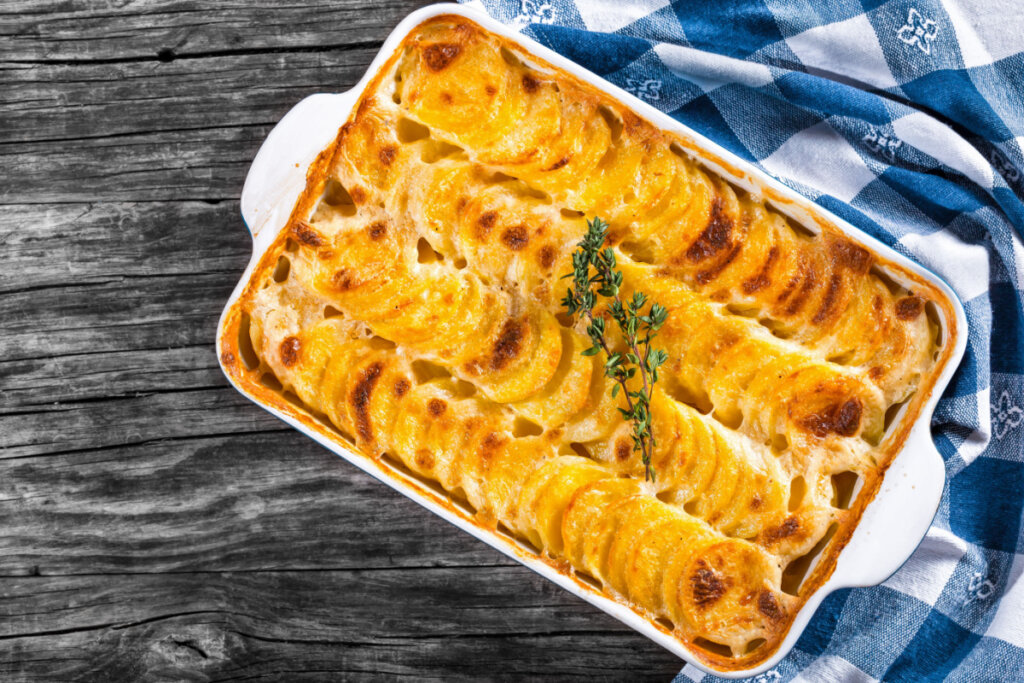 Batata gratinada em uma travessa branca sobre uma mesa de madeira. Ao lado, pano xadrez branco e azul 