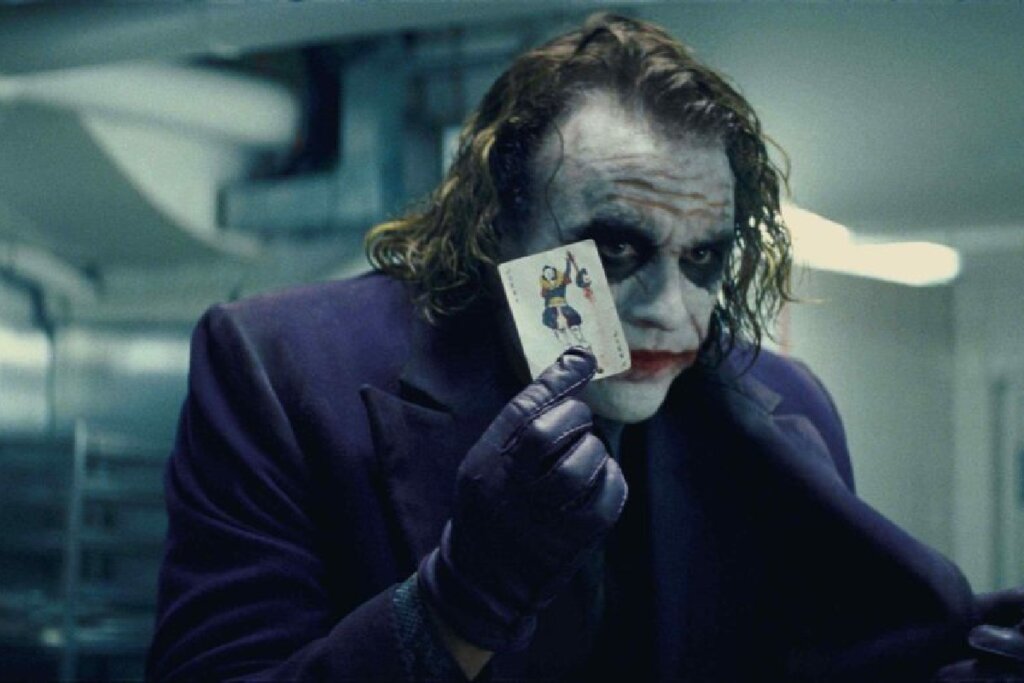 Um plano médio do vilão Coringa, com o rosto pintado de branco de forma irregular, sombras pretas pesadas ao redor dos olhos e um sorriso vermelho estendido por cicatrizes. Ele veste um paletó roxo e luvas de couro da mesma cor. O personagem segura uma carta de baralho (um curinga) entre os dedos da mão direita, posicionando-a à frente de seu rosto, enquanto olha fixamente para a câmera com uma expressão sinistra. O fundo é um ambiente interno clínico e levemente desfocado.
