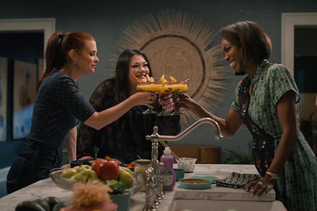 Três amigas sorridentes brindam com taças de margarita amarelas na bancada de uma cozinha moderna à noite.
