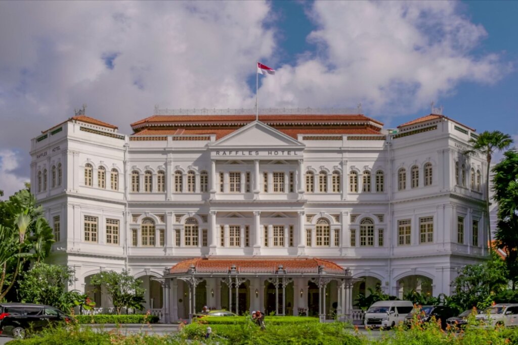 Fachada do Hotel Raffles com céu azul com nuvens acima e jardim verde na frente