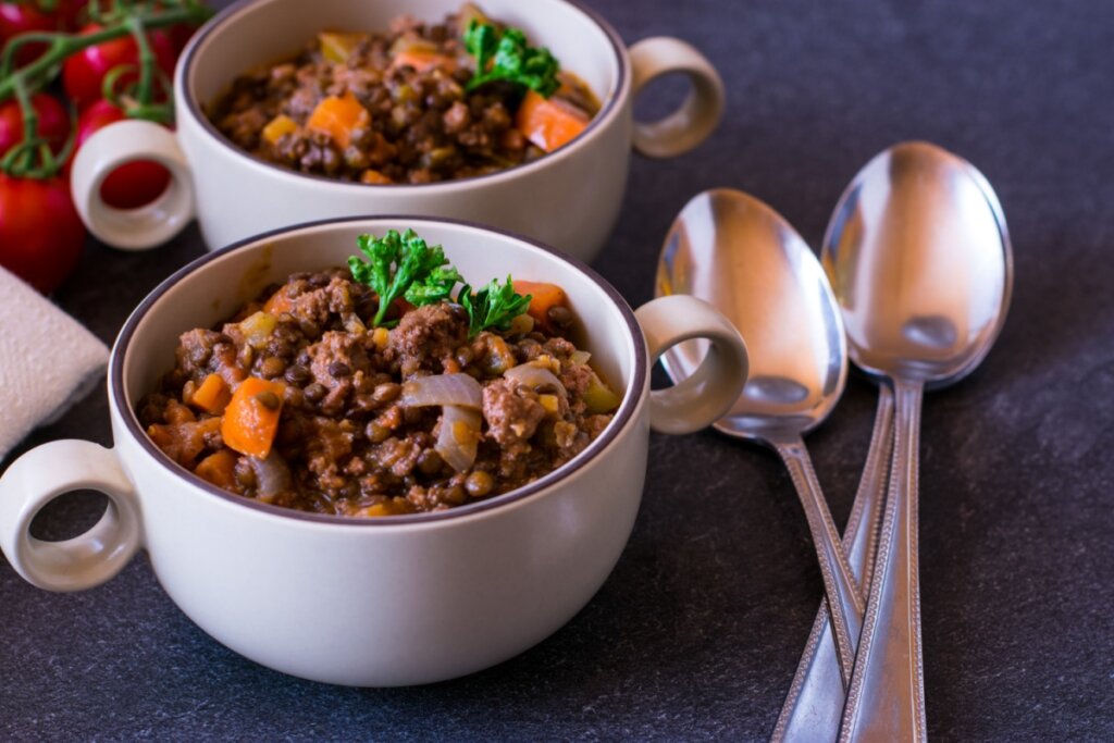 Duas tigelas de cerâmica clara com alças contêm um cozido encorpado de lentilhas com carne moída, pedaços de cenoura e cebola, decorado com folhas de salsa fresca. Três colheres de metal estão dispostas ao lado das tigelas sobre uma superfície escura. Ao fundo, à esquerda, aparecem alguns tomates na rama.
