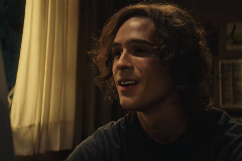 Um jovem de cabelos castanhos cacheados e compridos, iluminado por uma luz lateral quente que destaca seu rosto em um ambiente levemente obscurecido. Ele está com a boca entreaberta, sugerindo que está falando ou reagindo a algo em uma tela à sua frente (fora de campo). Ao fundo, nota-se uma cortina clara e quadros na parede em tons neutros.