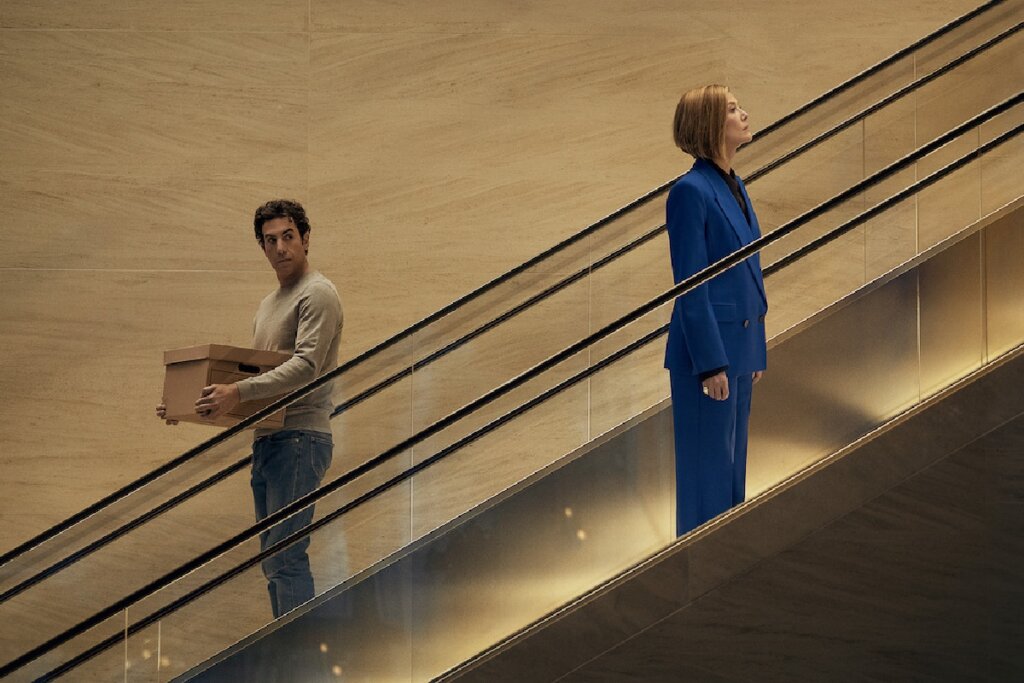 Fotografia de perfil em um ambiente moderno com paredes de mármore bege claro. Duas pessoas estão em escadas rolantes paralelas que se movem em direções opostas. À direita, uma mulher com cabelo chanel loiro-escuro veste um elegante terno azul-bic e olha fixamente para a frente, subindo. À esquerda, um homem de cabelos cacheados, vestindo blusa de moletom cinza e calça jeans, desce a escada enquanto segura uma caixa de arquivo de papelão e olha para trás, na direção da mulher.