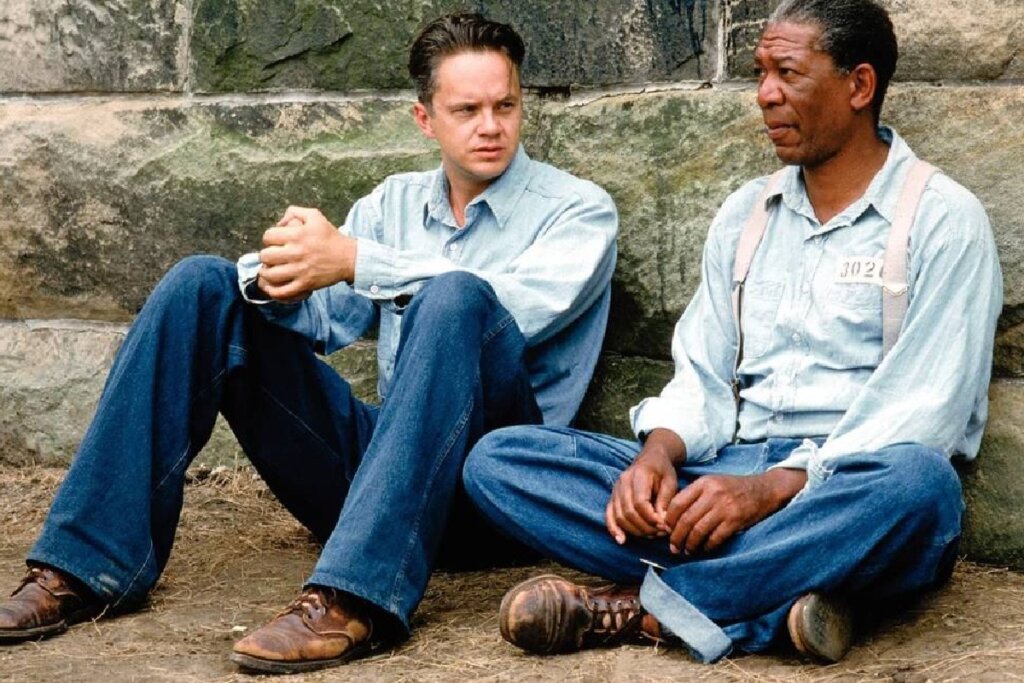 Dois homens estão sentados no chão, encostados em uma parede de pedras rústicas e claras em um pátio de prisão. À esquerda, um homem branco (Andy Dufresne) veste uma camisa de brim azul claro de mangas longas e calças jeans escuras, com as mãos entrelaçadas sobre os joelhos. À direita, um homem negro (Ellis "Red" Redding) usa uma camisa semelhante com suspensórios claros e um crachá de identificação prisional, olhando atentamente para o companheiro enquanto conversam.