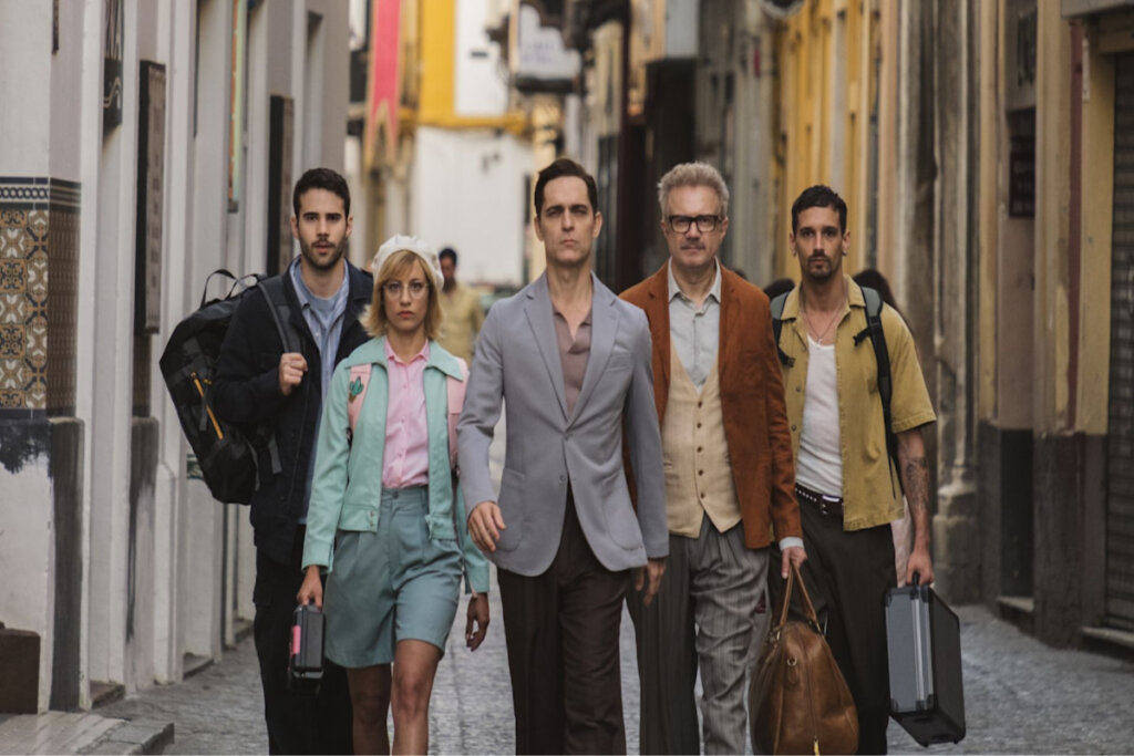 A imagem mostra cinco pessoas caminhando por uma rua estreita de aparência europeia, carregando malas e equipamentos. O grupo tem expressões sérias e postura determinada, transmitindo a sensação de uma missão ou plano em andamento
