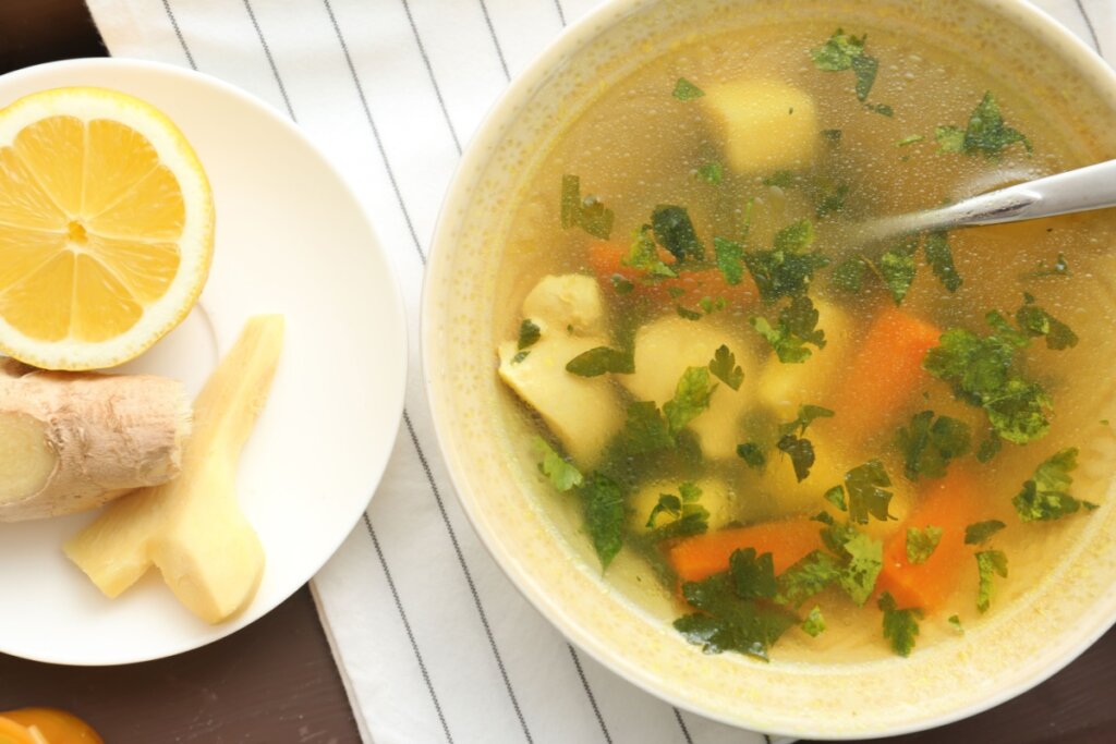 Sopa de legumes com gengibre servido em tigela de cerâmica branca em cima de toalha de mesa listrada em preto e branco em cima de mesa de madeira, ao lado um prato com um pedaço de gengibre e rodela de limão siciliano