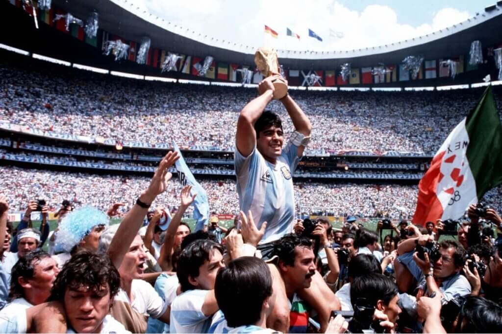 Uma imagem de corpo inteiro de Diego Maradona, um dos maiores jogadores de futebol de todos os tempos, levantando a Taça da Copa do Mundo. Ele está em cima dos ombros de um torcedor, com a multidão e os jogadores da Seleção Argentina ao seu redor. O fundo é um estádio de futebol.