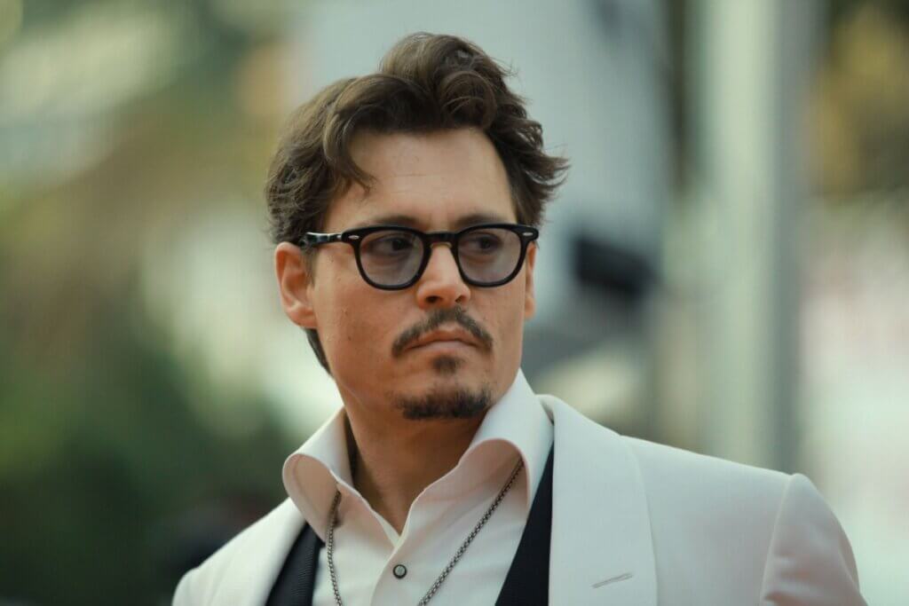 Fotografia em plano médio do ator Johnny Depp olhando de perfil parcial para a direita com uma expressão séria. Ele usa óculos de grau com armação preta, terno com paletó branco, colete preto e camisa social branca com o colarinho aberto.