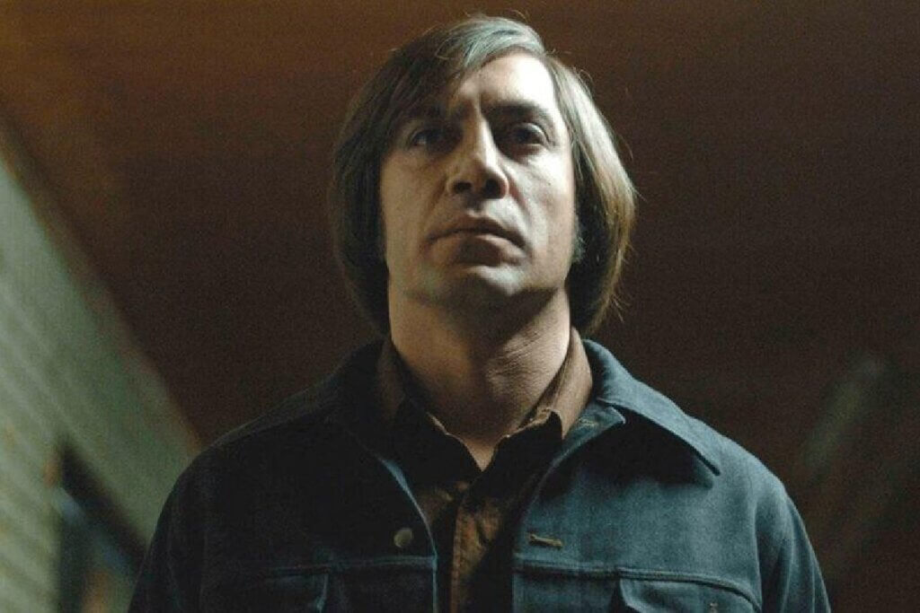 Close-up em ângulo de baixo para cima do personagem Anton Chigurh. Ele tem uma expressão facial completamente séria, fria e impassível, com um corte de cabelo estilo chanel rústico com franja. Veste uma jaqueta escura abotoada sobre uma camisa marrom. A iluminação vem de cima, destacando suas feições contra um fundo escuro e sutilmente desfocado.