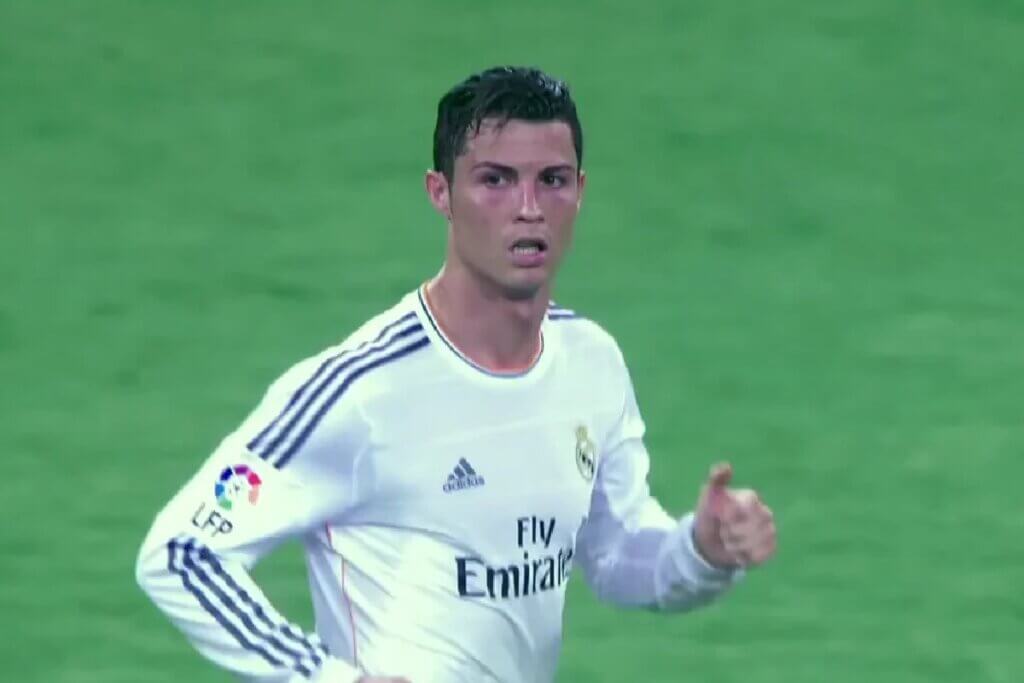 Uma imagem de rosto de Cristiano Ronaldo, um dos melhores jogadores de futebol de todos os tempos, com a camisa branca e azul do Real Madrid, o uniforme número sete. Ele está olhando para a câmera com uma expressão séria. O fundo é um campo de futebol.