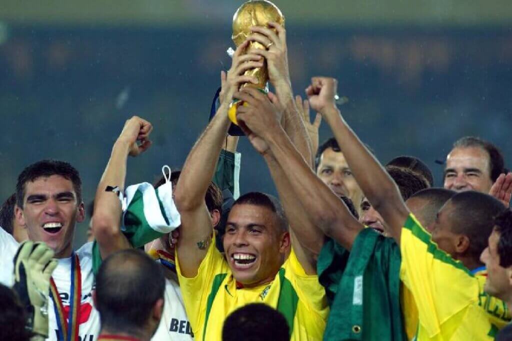 Uma imagem de corpo inteiro de Ronaldo Nazário, segurando a Taça da Copa do Mundo. Ele está sorrindo para a câmera, com a medalha de campeão ao redor do pescoço. O fundo é uma multidão borrada em um estádio.