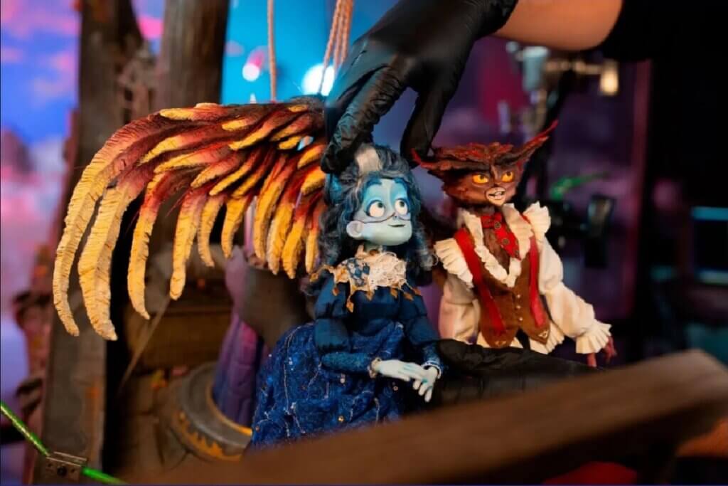 astidores de uma produção em stop-motion. Uma mão usando luva cirúrgica preta ajusta delicadamente a cabeça de uma boneca com pele azul claro, cabelos escuros e vestido azul imperial de época. Ao lado dela está outro boneco com feições de coruja vestindo colete marrom. O cenário de fundo é desfocado.