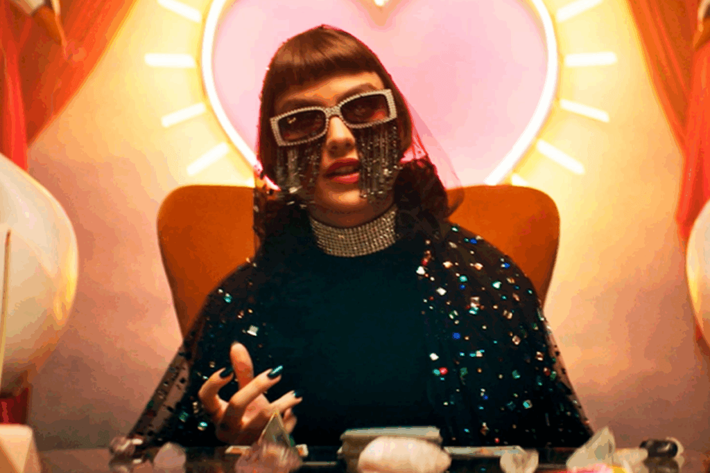 Mulher usando óculos decorados e roupas brilhantes, sentada diante de uma mesa com cristais sob uma iluminação colorida em formato de coração