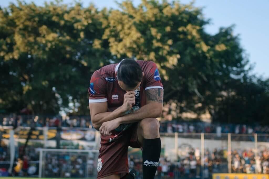 Um jogador de futebol de perfil, ajoelhado no gramado de um campo. Ele veste um uniforme esportivo nas cores marrom e vinho com detalhes brancos, além de meiões pretos. O atleta está com o tronco inclinado, a cabeça baixa e leva uma das mãos ao rosto. O fundo mostra a linha do horizonte de um campo de várzea, com árvores frondosas iluminadas pelo sol e uma arquibancada com público ao longe.