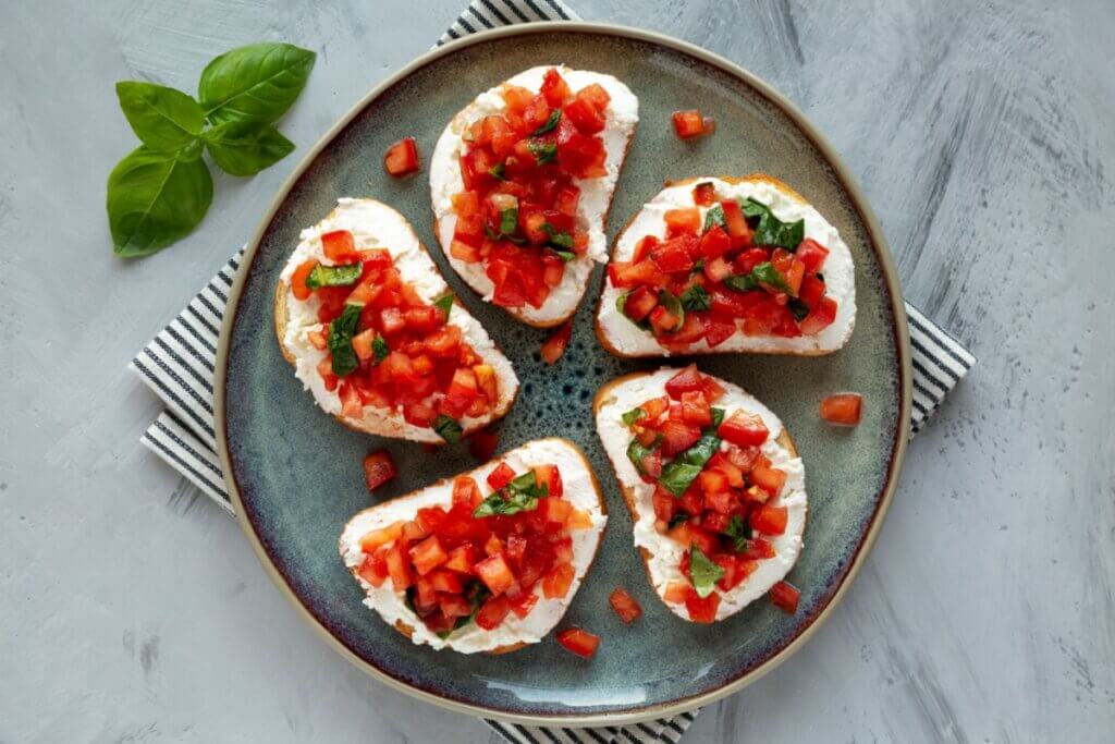 Bruschettas com ricota, tomate em cubos e folhas de manjericão servidas em fatias de pão sobre um prato redondo