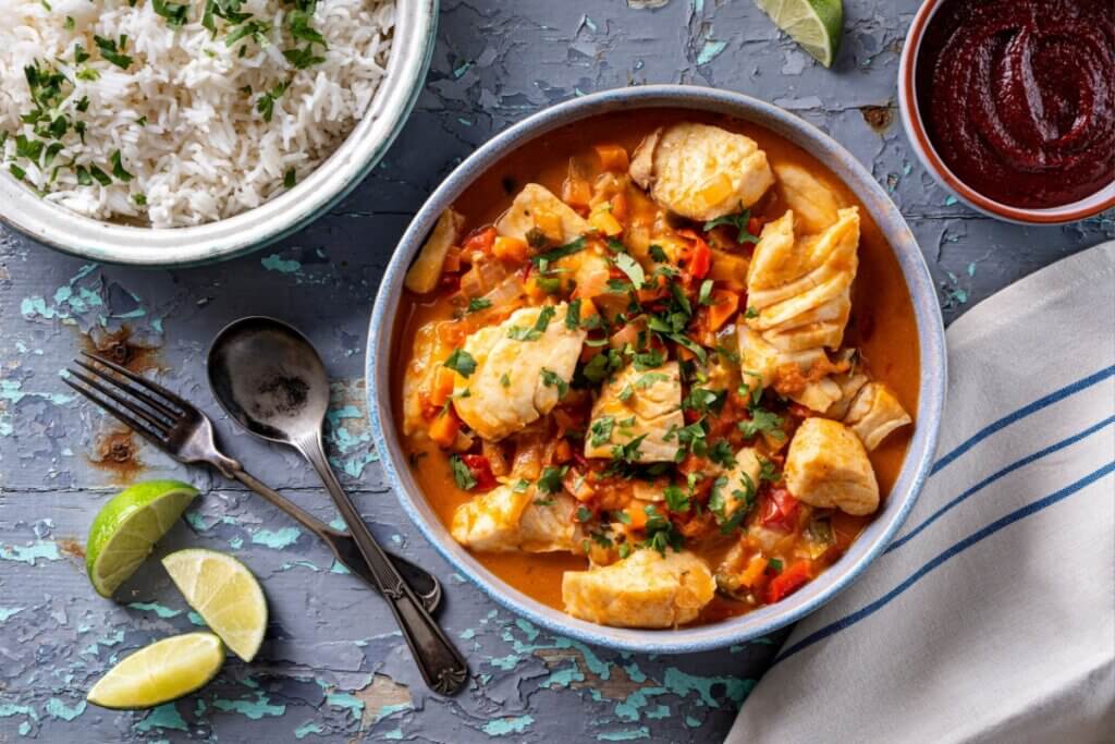 Uma Moqueca de Peixe vibrante, com pedaços suculentos de peixe branco em um molho alaranjado de tomates e pimentões, finalizada com coentro fresco. Uma tigela de arroz branco soltinho e um pequeno bowl com molho de pimenta (ou pirão) bem avermelhado.