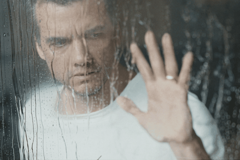 Um homem de camiseta branca olha para fora através de um vidro molhado pela chuva, com a mão espalmada contra a superfície, onde se vê uma aliança.