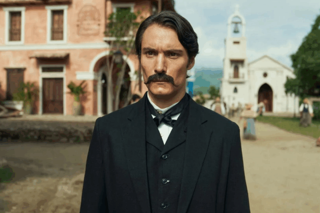 Um homem de bigode proeminente veste um terno preto com gravata borboleta e olha para a câmera, com prédios de estilo colonial e uma torre de igreja ao fundo.