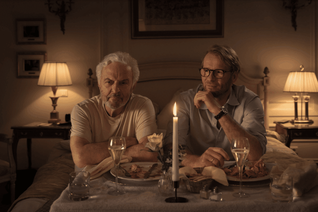 Dois homens sentados diante de uma mesa posta com taças e vela acesa em um quarto.