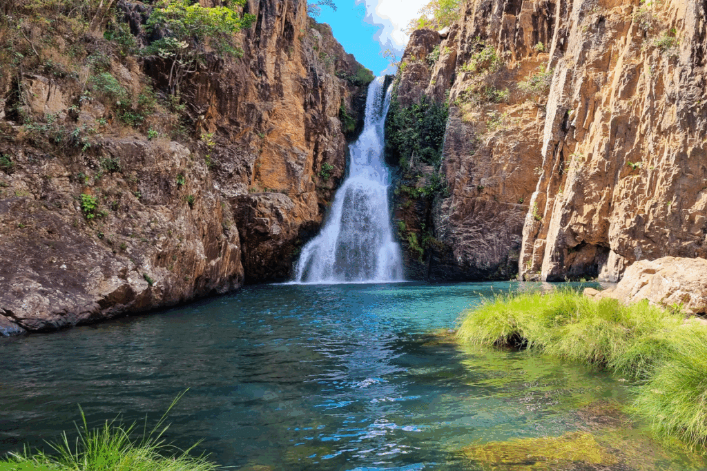 Cachoeira caindo entre paredões de rocha avermelhada sobre um poço de águas azul-esverdeadas, cercado por vegetação verde na Chapada dos Veadeiros.