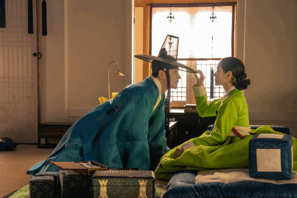 Imagem de um casal em trajes tradicionais coreanos, em um ambiente interno, com o homem usando roupas azuis e chapéu tradicional preto e a mulher vestindo um traje verde vibrante. Os dois estão próximos, em uma cena de interação.