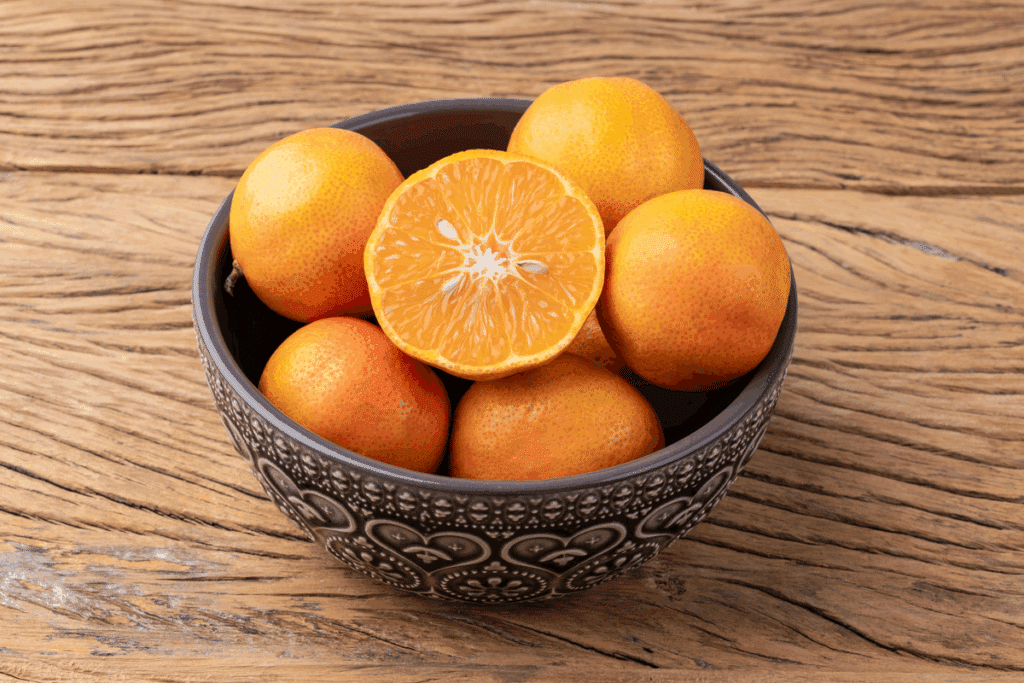 Tigela com fruta laranja inteira e uma metade cortada mostrando a polpa suculenta, sobre mesa de madeira