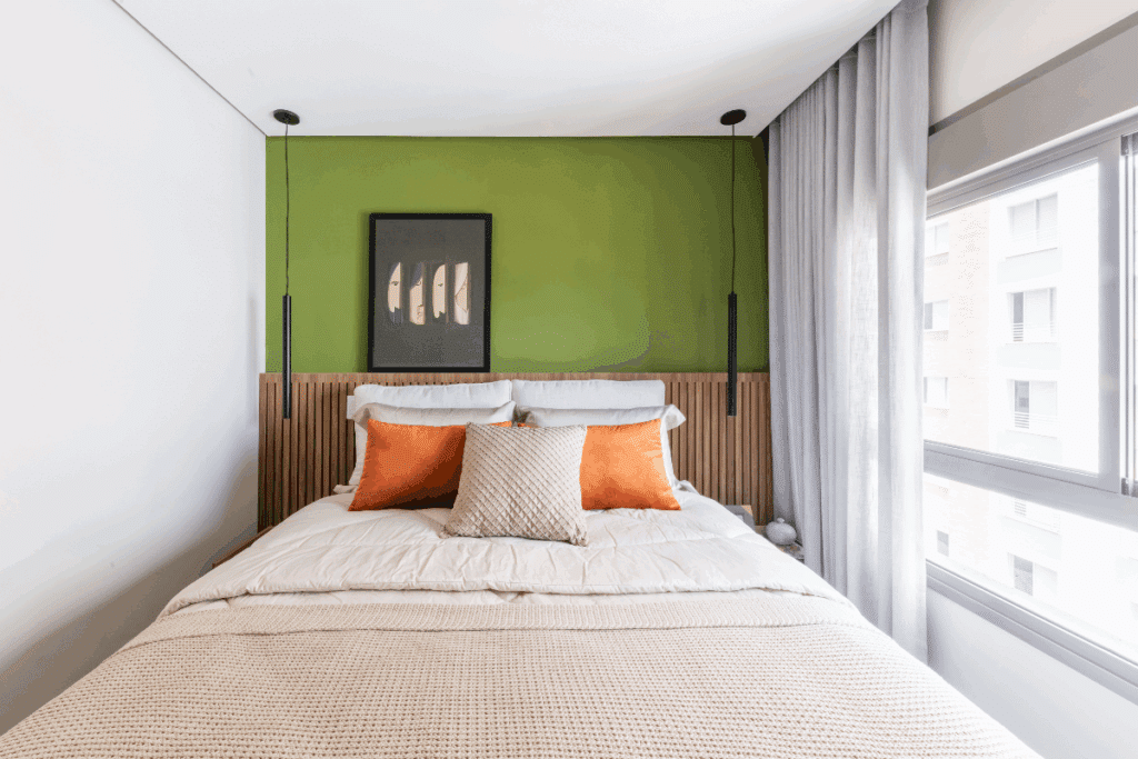 Um quarto moderno e aconchegante, com cama de casal revestida por roupas de cama claras e almofadas em tons de laranja e bege. A parede verde atrás da cama contrasta com a cabeceira de madeira ripada e recebe um quadro decorativo. Duas luminárias pendentes ficam nas laterais, enquanto uma ampla janela com cortinas cinza permite a entrada de bastante luz natural.