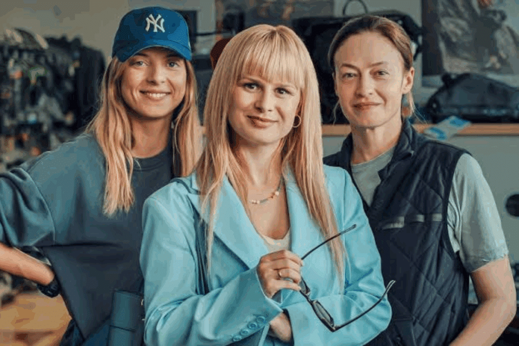 Três mulheres sorriem lado a lado. A mulher da esquerda usa boné azul, a do meio usa um terno azul claro e segura óculos, e a da direita veste um colete escuro.