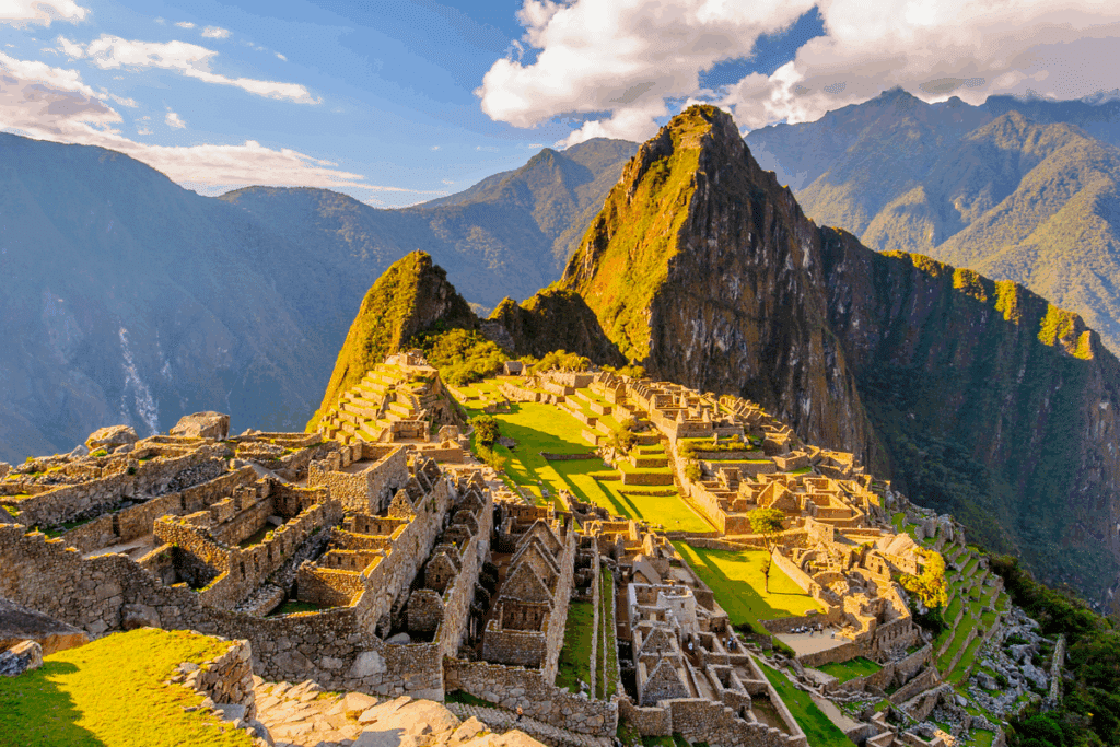 Fotografia panorâmica da cidadela inca de Machu Picchu, no Peru, com ruínas de construções de pedra, terraços agrícolas e gramados iluminados pelo sol. Ao fundo, a montanha Huayna Picchu, pontiaguda e coberta de vegetação, cercada por montanhas verdes e nuvens brancas em um céu azul.